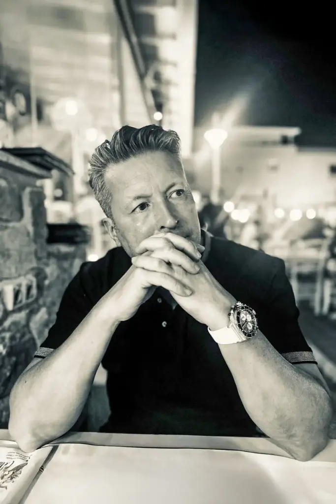 Entspanntes Schwarz-Weiß-Porträt von Zahntechniker Kes Carpenter, der nachdenklich mit verschränkten Händen an einem Tisch sitzt.