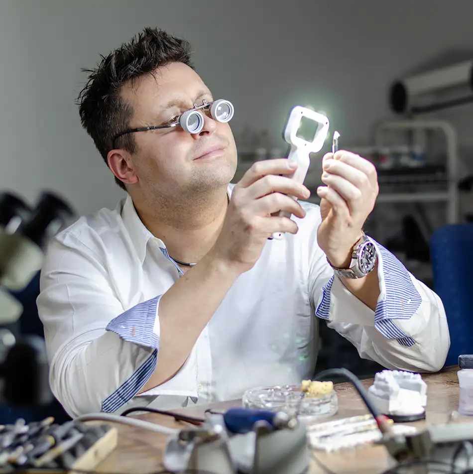 Zahntechniker Kes Carpenter prüft am Werktisch im Dentallabor in Hennigsdorf mit einer beleuchteten Lupe konzentriert ein zahntechnisches Werkstück.